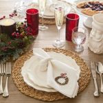 Christmas dinner table setting with a woven charger mat and a stack of white layered dinner plates. There are red water cups on the table as well as champagne flutes, silver utensils, and a christmas wreath