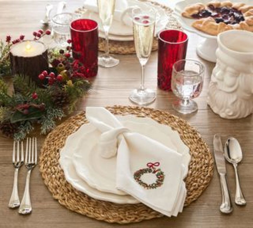 Christmas dinner table setting with a woven charger mat and a stack of white layered dinner plates. There are red water cups on the table as well as champagne flutes, silver utensils, and a christmas wreath