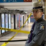 A New York State police officer wearing glasses, a hat, and a gray uniform stands guard outside of a Penn Station hallway cordoned off with caution tape.