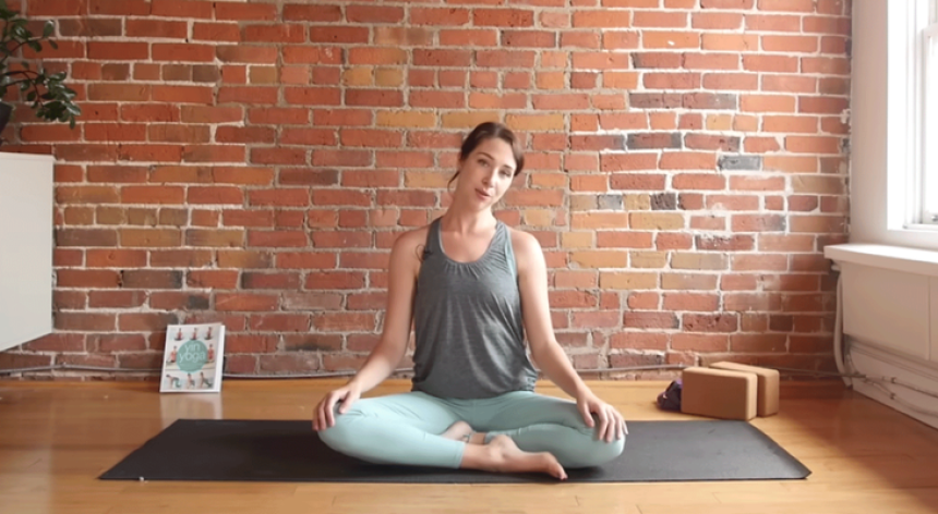 Kassandra Reinhardt teaching a 10-minute morning yoga session while seated and tilting her head and neck to one side during a full-body stretching routine