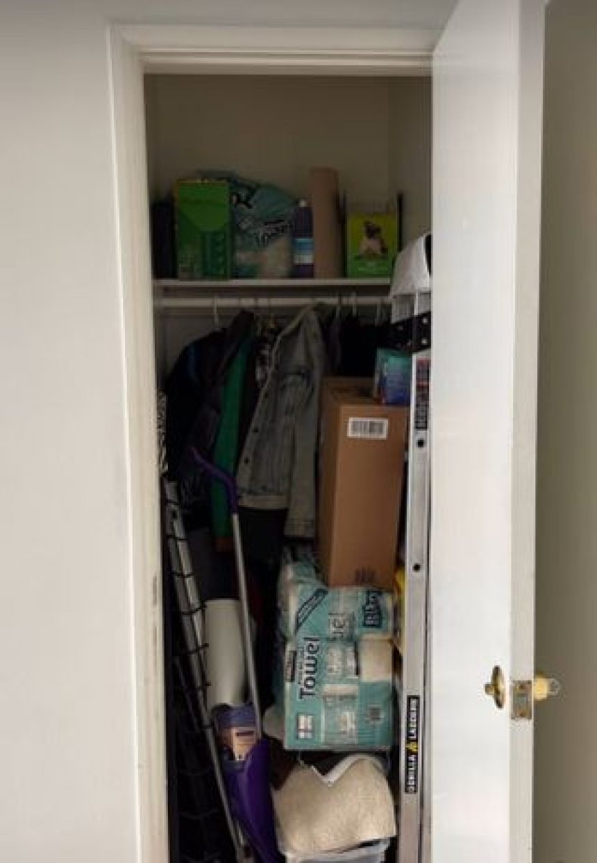 Image of an open small closet that is full of cleaning supplies, jackets, toilet paper, and a mix of random house items.