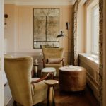 Backed by an antique glass mirror, two velvet upholstered wingback chairs sit in front of a window.