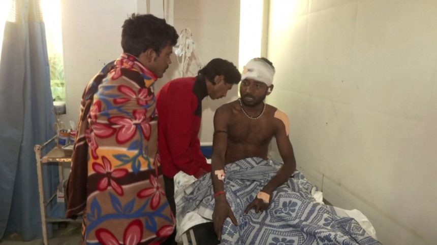 An injured man sits in a hospital bed with a thick bandage around his head as he is examined by a doctor.