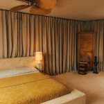 A beige, brown, and yellow bedroom. The bed is a textured velvet bed with mustard yellow bedding. Along the back wall is a thick line of taupe curtains.