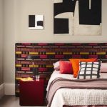 A white bedroom with bed that has a colorful brown, yellow, and pink patterned, extended headboard. There is a maroon, cube bedside table and a dark red rug in the room as well