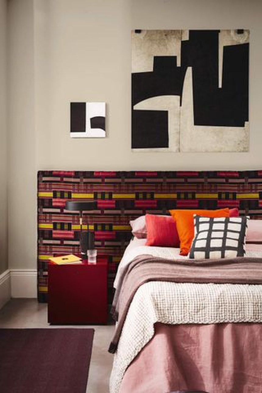 A white bedroom with bed that has a colorful brown, yellow, and pink patterned, extended headboard. There is a maroon, cube bedside table and a dark red rug in the room as well