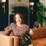 A portrait of a woman sitting on a leather couch. She holds an emmy award