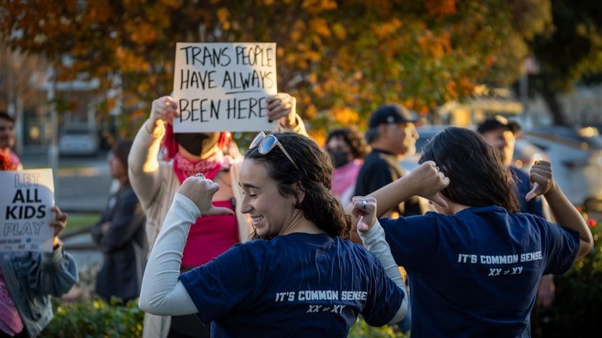 Transgender athlete supporters hold up signs at left as Tori Hitchcock, center, of the Young Women for America, and Salomay McCullough, right, both former female athletes, show off their "Save Girls Sports" shirts as an overflow crowd converges outside the Riverside Unified School District meeting Thursday night to debate the rights of transgender athletes to compete in high school sports Thursday, Dec. 19, 2024. 