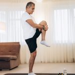 A man standing on one leg and stretching another in a living room next to a yoga mat and some weights