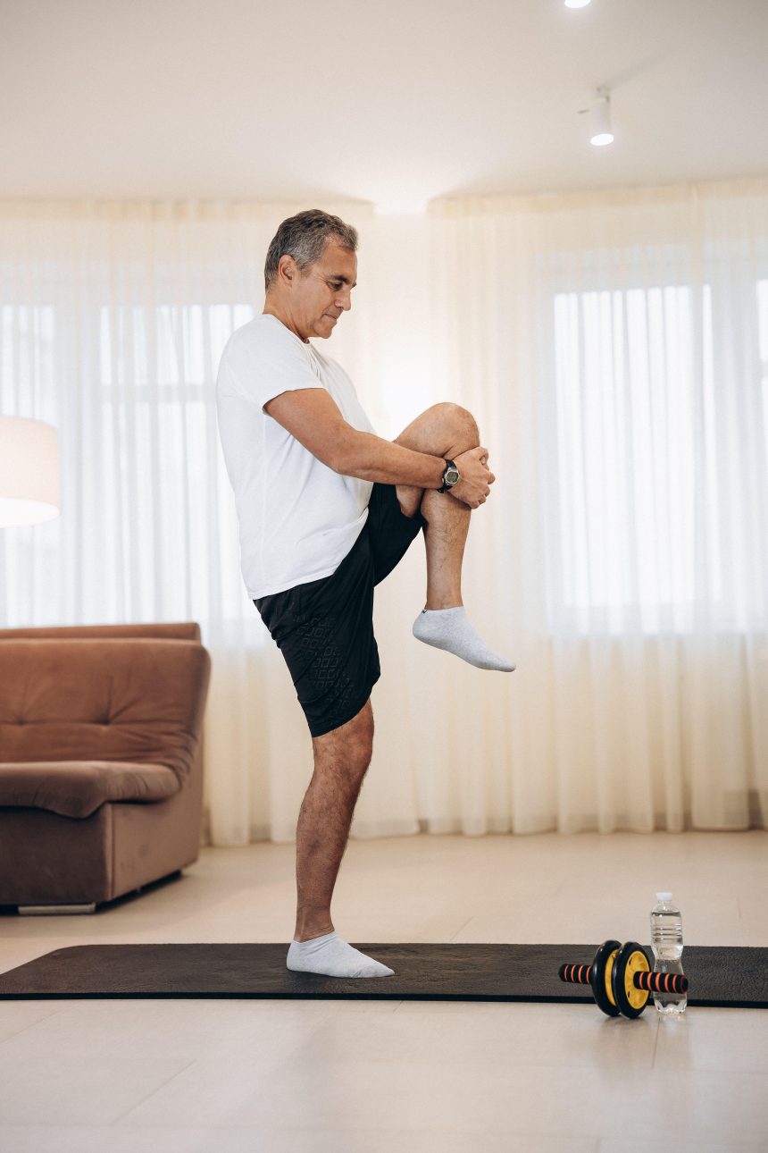 A man standing on one leg and stretching another in a living room next to a yoga mat and some weights