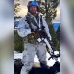 Matthew Livelsberger wearing cold weather gear and holding a rifle while standing over a snowmobile