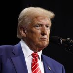 Trump speaks behind a microphone wearing a blue suit, white shirt and red tie