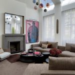 modern living room with fireplace with large artwork hung on either side and a mirror hanging over the fire, two windows with sheer curtains, a large L-shaped brown sofa with a nesting coffee table and modern white and timber armchair