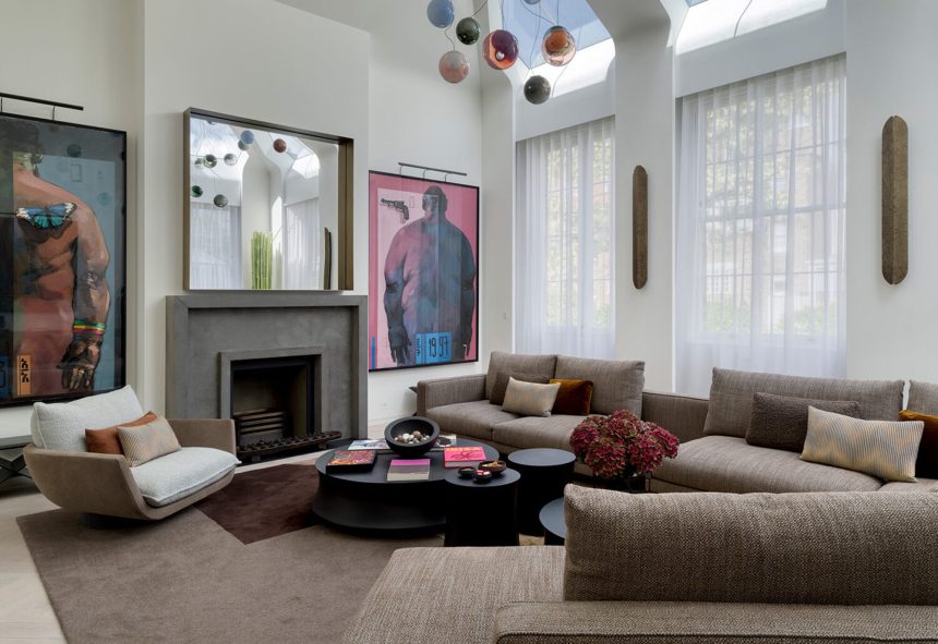 modern living room with fireplace with large artwork hung on either side and a mirror hanging over the fire, two windows with sheer curtains, a large L-shaped brown sofa with a nesting coffee table and modern white and timber armchair