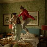 Image of a woman and two children jumping on an unmade bed. The headboard is blue and there are two orange lamps on either side of the bed. The room has green floral wallpaper all around it.