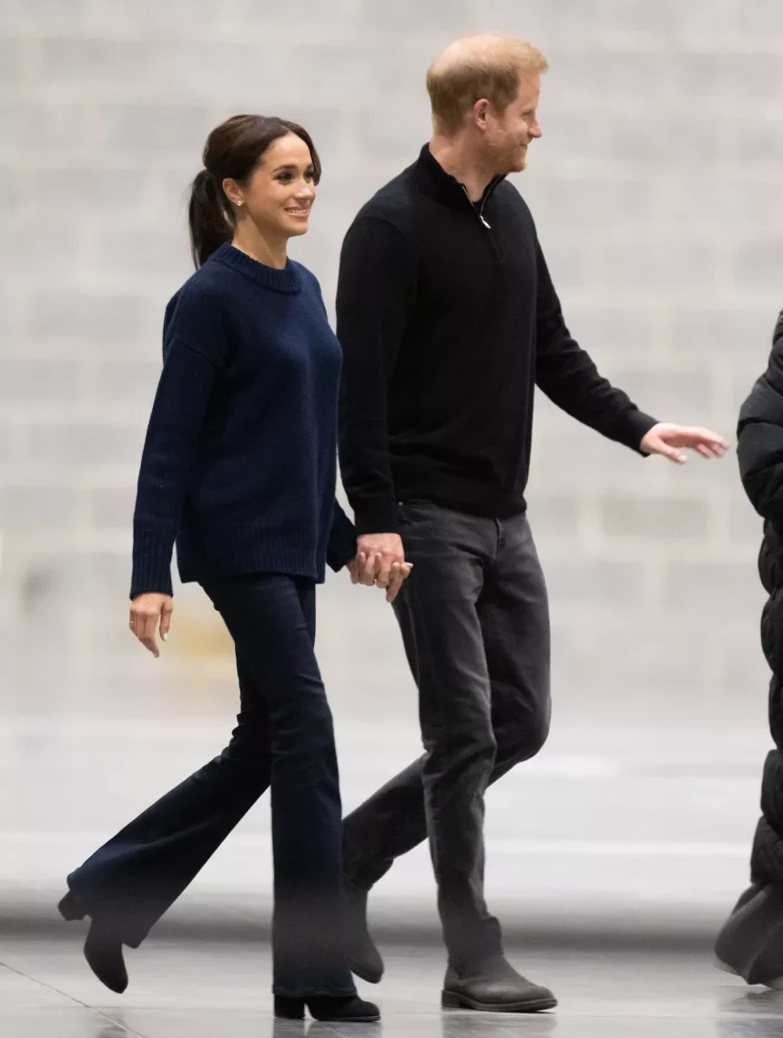 Meghan Markle wears a navy sweater, black bootcut jeans, and black boots while walking with Prince Harry