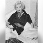 A black and white photo of Ruth Ellis on her white bed posing for the camera.