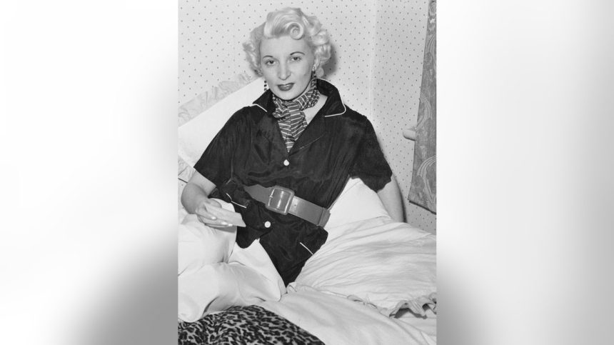 A black and white photo of Ruth Ellis on her white bed posing for the camera.