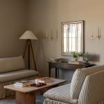 A beige living room with two armless love seats that are facing across from one another. There is a natural wood coffee table between them. In the corner is a wood floor lamp with a white fabric shade.