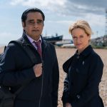 Two detectives standing on beach looking into camera