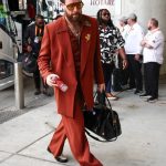 Travis Kelce struts into the stadium in a burnt orange suit with a sparkly shirt, oversized sunglasses, and a gold floral brooch, carrying a black leather bag while holding a can of drink.