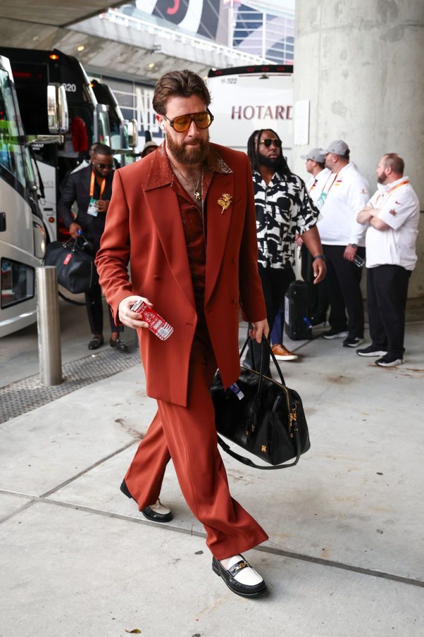 Travis Kelce struts into the stadium in a burnt orange suit with a sparkly shirt, oversized sunglasses, and a gold floral brooch, carrying a black leather bag while holding a can of drink.