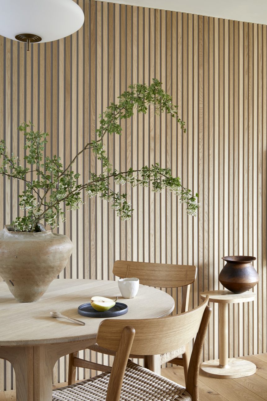 Natural woods and textures used in a home. On the dining table is a neutral toned plant planter with green foliage hanging out of it. On the right side of the room is a small side table with a large accessory on top.